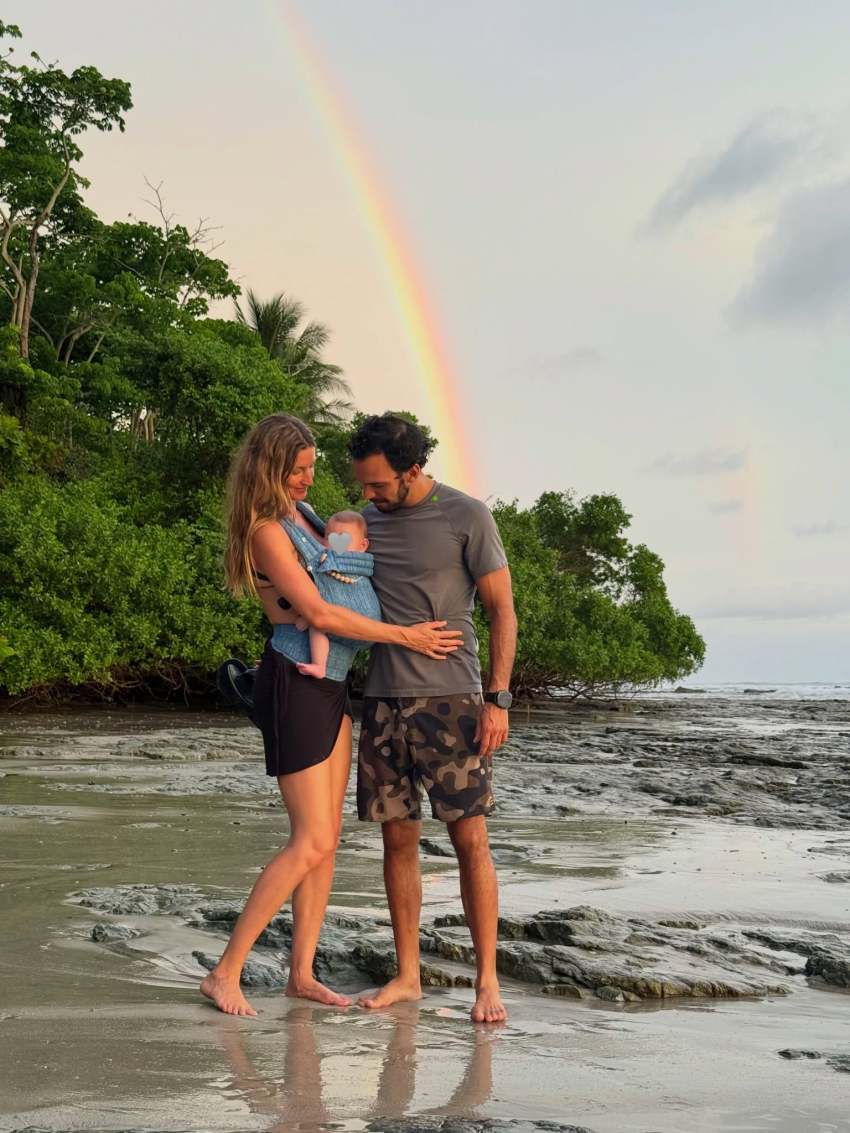 A woman in a black swimsuit holding a baby in a carrier and a man in camo shorts stand together on a rocky beach with a rainbow in the sky behind them.