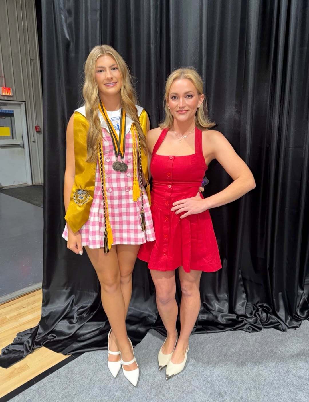 Two blonde women stand together in front of a black curtain; the taller one wears a gold graduation gown with medals and a pink plaid dress, the other wears a red dress.
