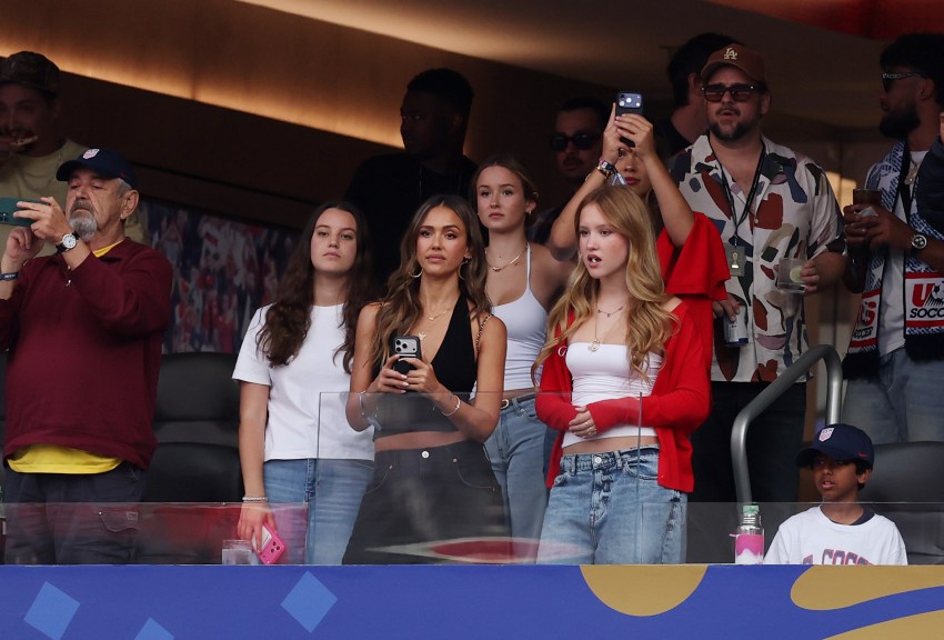 A woman in a black halter top stands with two teen girls in a stadium box, holding a phone and watching a soccer match.