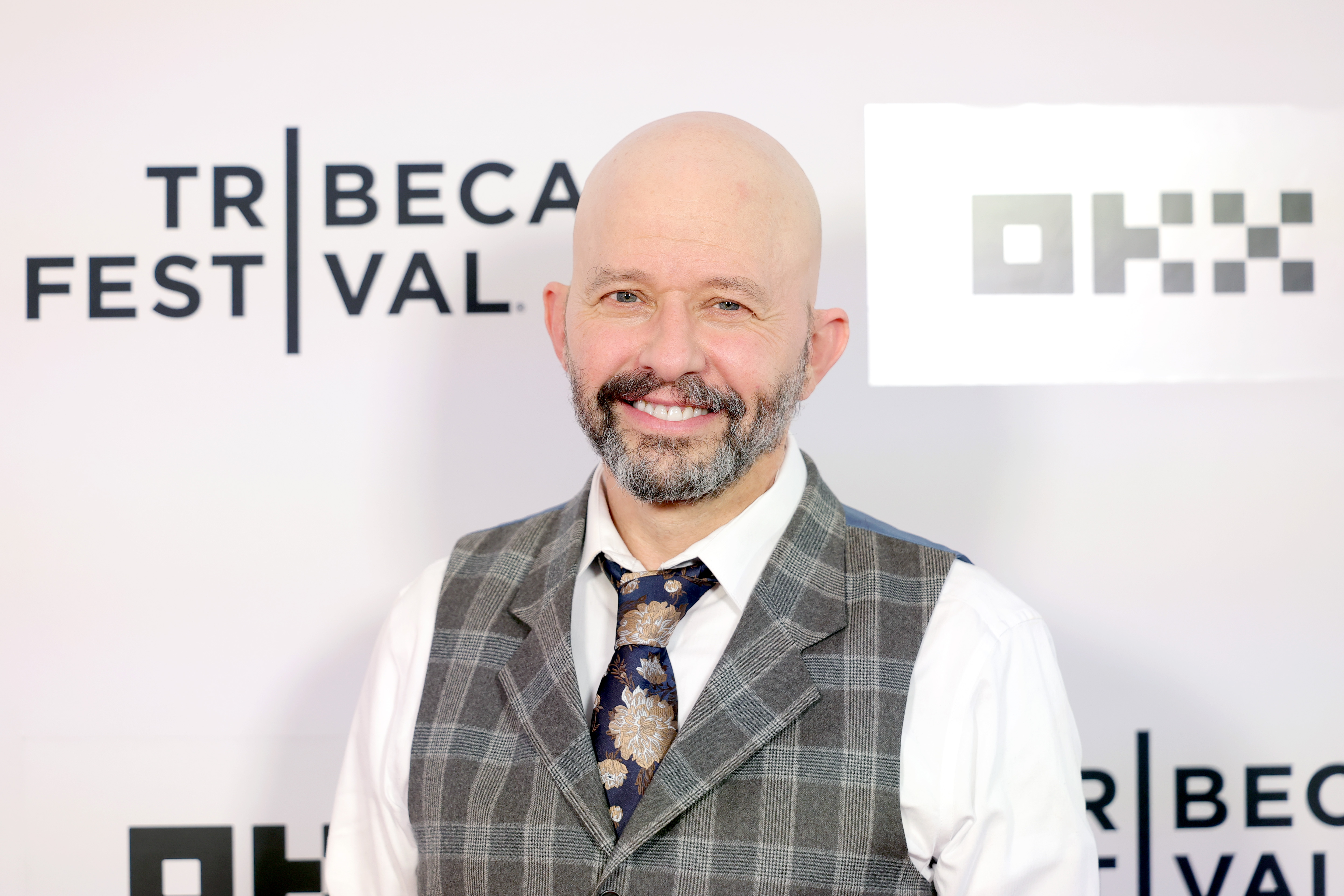Click to open the image gallery Bald man with a gray goatee wearing a plaid vest and floral tie smiling in front of a Tribeca Festival step-and-repeat backdrop.