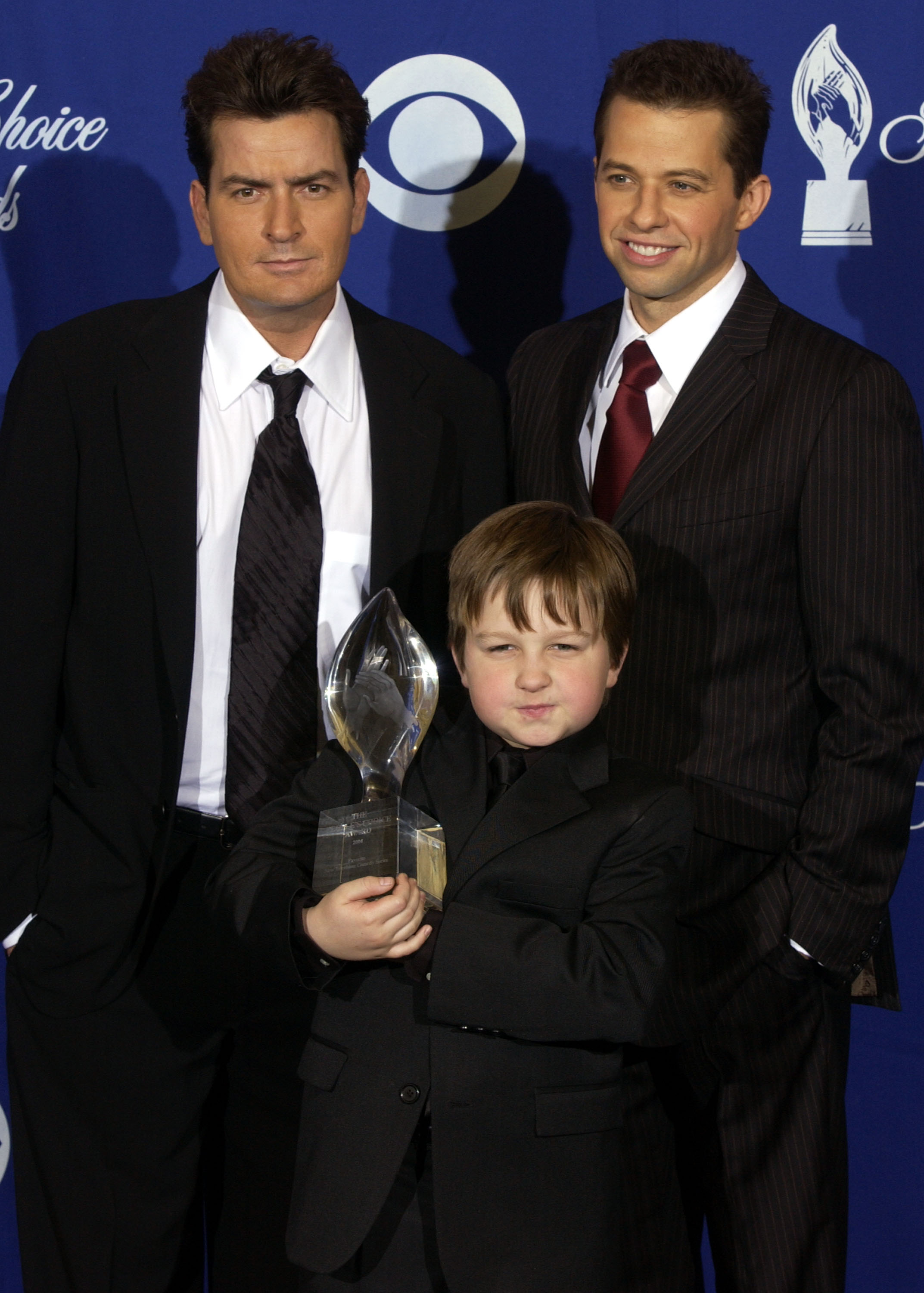 Click to open the image gallery Two adult men in dark suits standing behind a young boy in a black suit holding a crystal trophy at the People's Choice Awards pressroom.