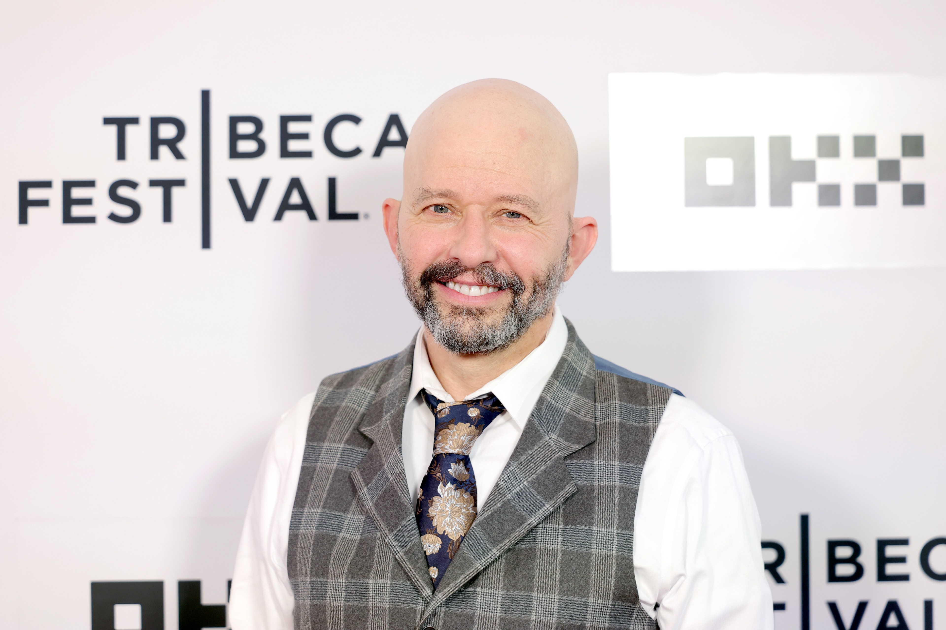 Bald man with a salt-and-pepper beard wearing a gray plaid vest and floral tie smiles in front of a Tribeca Festival step-and-repeat backdrop.