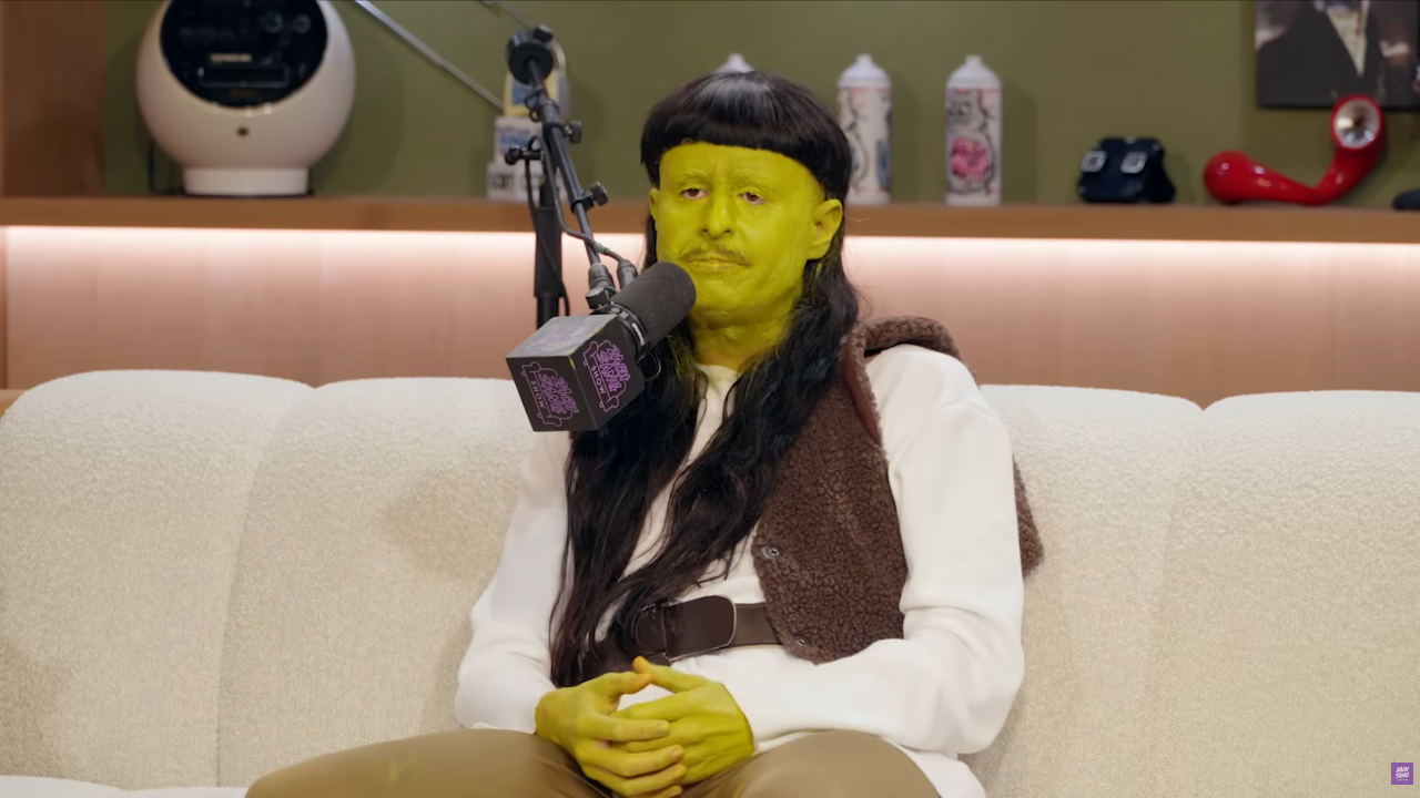 Person wearing green face paint, a long dark wig, and a brown vest sits on a white couch next to a podcast microphone.