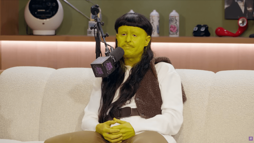 Person wearing green face paint, a long dark wig, and a brown vest sits on a white couch next to a podcast microphone.