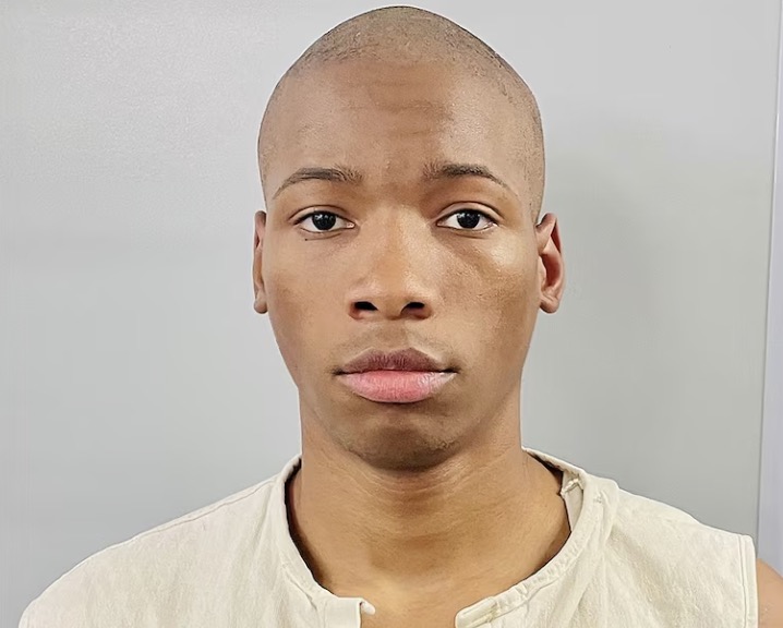 Booking photo of a young man with a shaved head wearing a beige sleeveless shirt against a gray background.