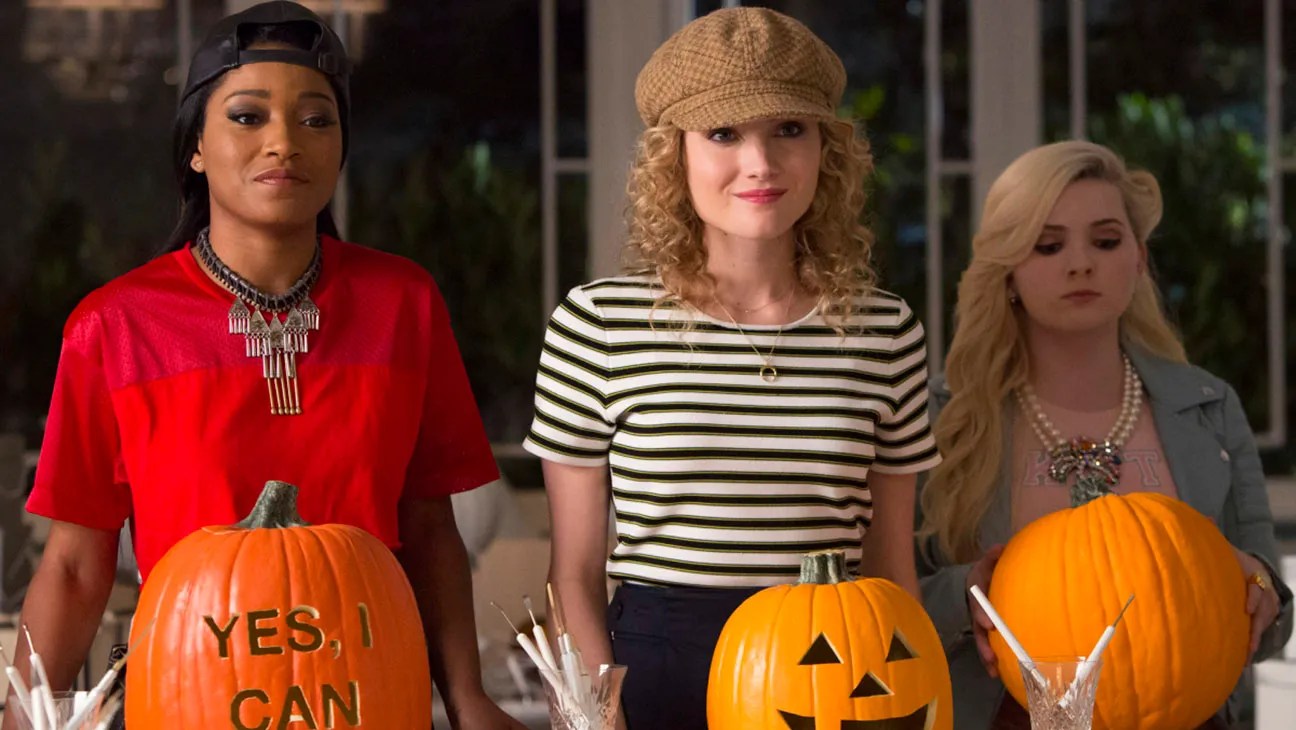 Three women standing behind Halloween pumpkins, one carved with a jack-o'-lantern face and one reading 'YES, I CAN,' in a TV scene.