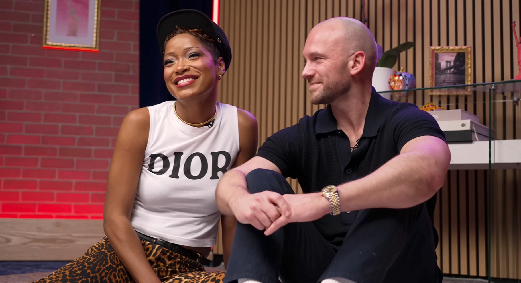 Keke Palmer in a white Dior tank top and backwards cap sits beside a bald man in a black polo shirt on a TV studio set.