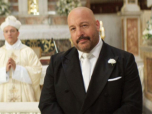 Bald man with beard in black tuxedo and white boutonniere standing inside an ornate church, with a robed altar server visible behind him.