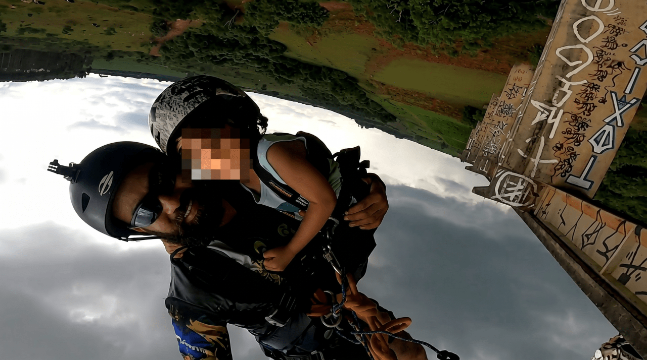 Upside-down GoPro shot of a helmeted man harnessed to a small child jumping from a graffiti-covered concrete bridge, cloudy sky below.