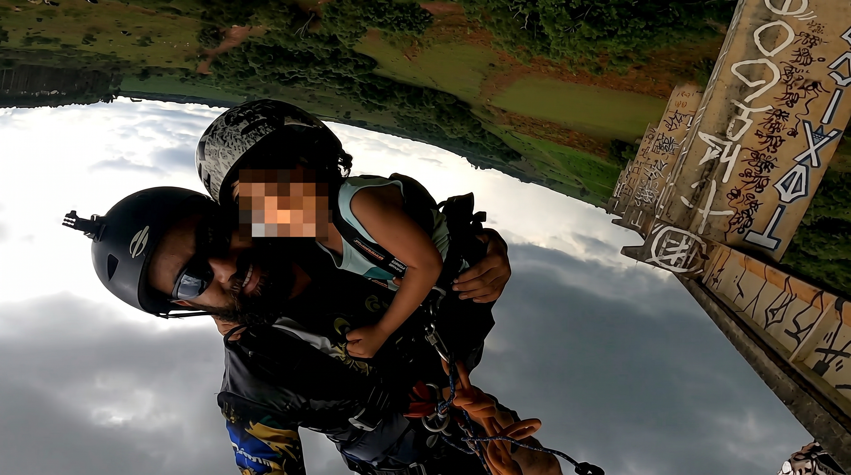 Upside-down GoPro shot of a helmeted man harnessed to a small child jumping from a graffiti-covered concrete bridge, cloudy sky below.