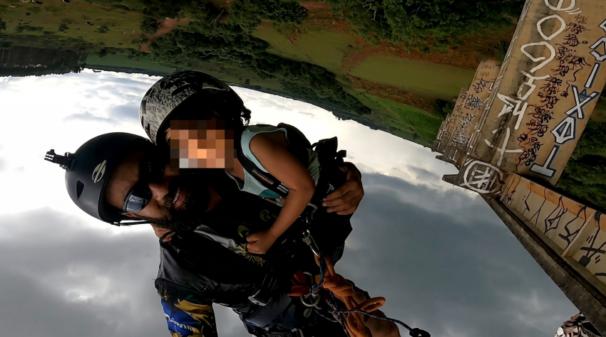 Upside-down GoPro shot of a helmeted man harnessed to a small child jumping from a graffiti-covered concrete bridge, cloudy sky below.