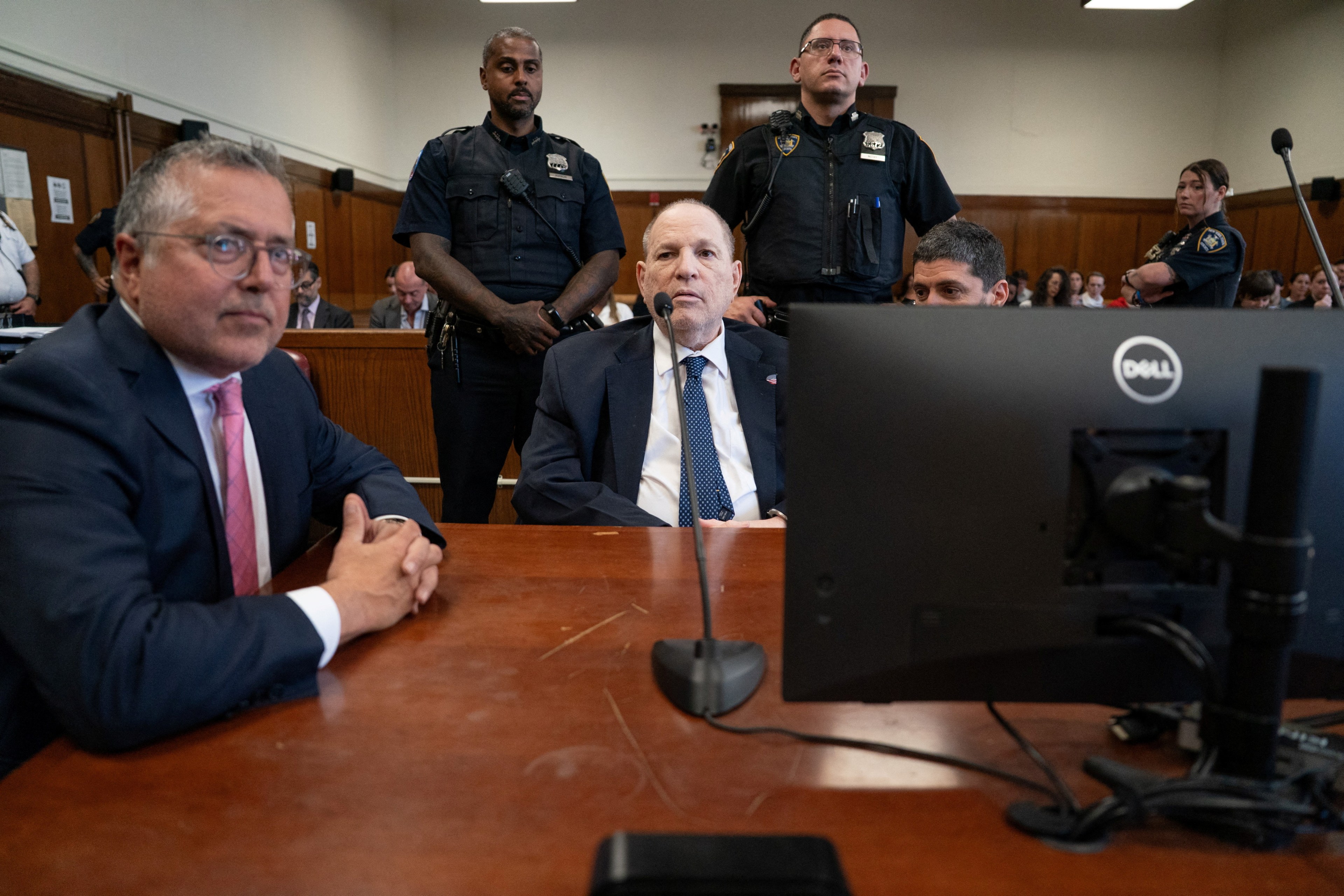Harvey Weinstein in a dark suit and blue tie sits at the defense table in a courtroom, flanked by his attorney and two uniformed court officers standing behind.