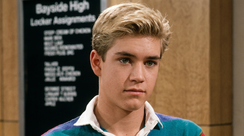 Young blonde man in a colorful striped polo shirt standing in front of a Bayside High locker assignments sign.