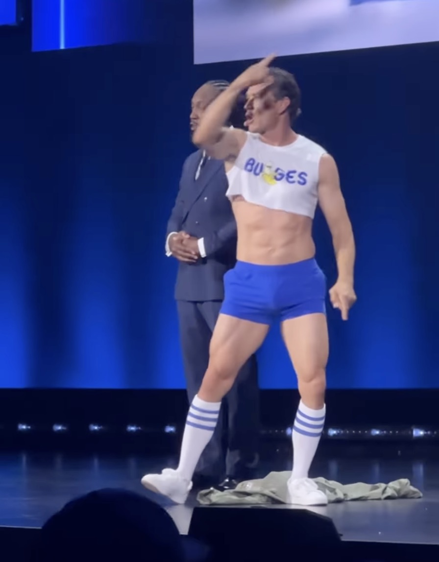 Man in white crop top and blue shorts posing on stage next to a man in a dark suit, with blue stage lighting behind them.