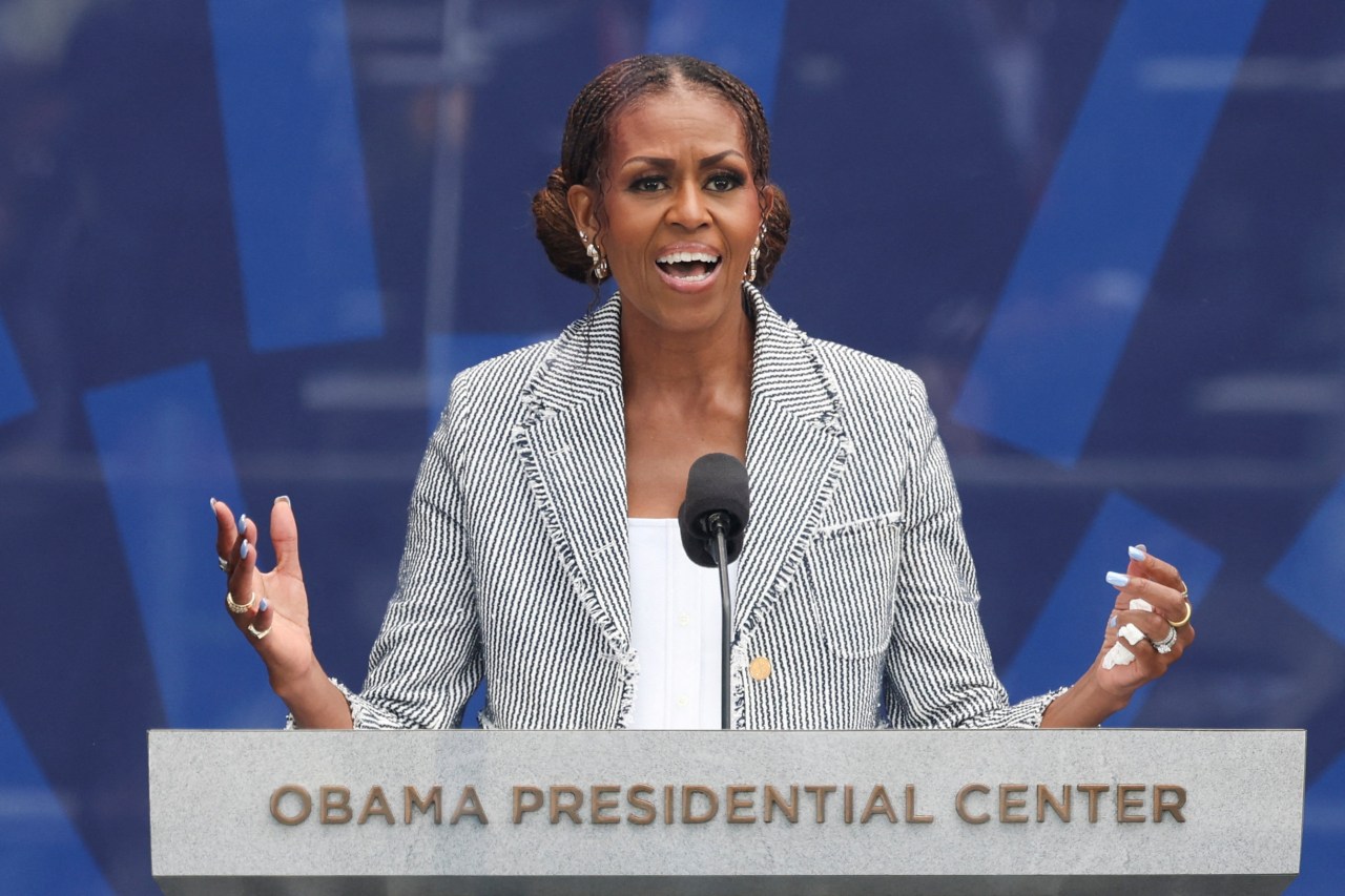 Michelle Obama speaking at a podium labeled 'Obama Presidential Center,' gesturing with both hands, wearing a striped blazer.