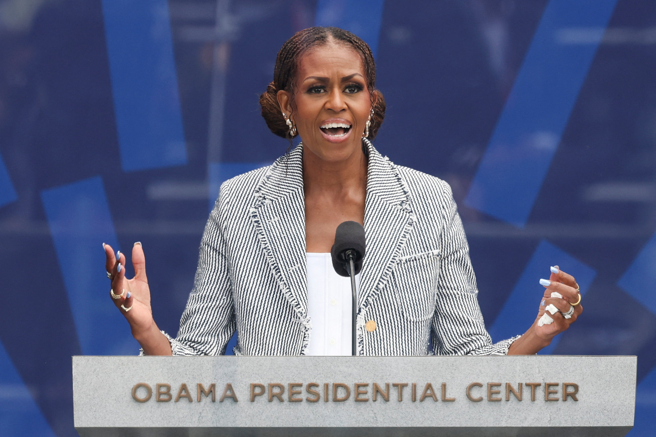 Michelle Obama speaking at a podium labeled 'Obama Presidential Center,' gesturing with both hands, wearing a striped blazer.