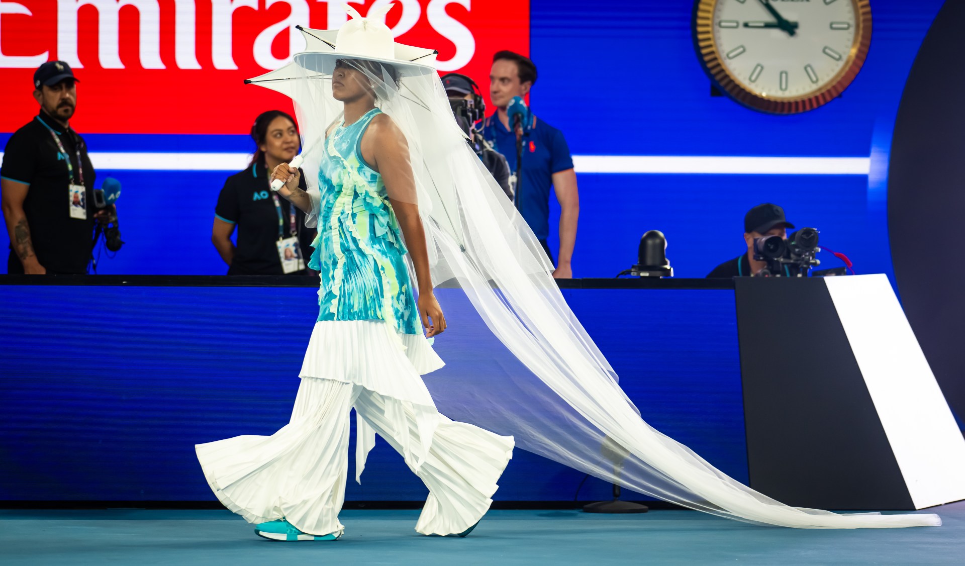 Tennis player in a white pleated outfit with a wide-brimmed hat and long sheer veil walks onto a blue court at the Australian Open.