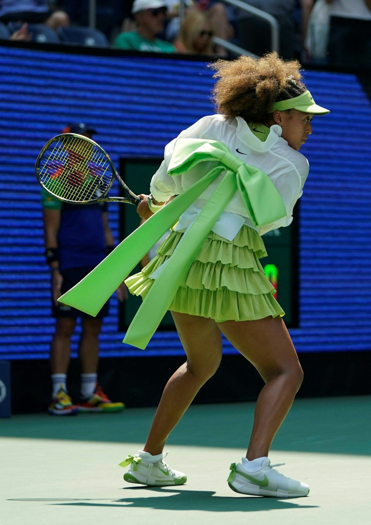 Tennis player from behind wearing a ruffled green skirt and white Nike jacket with a large lime-green bow tied at the back, holding a tennis racket.