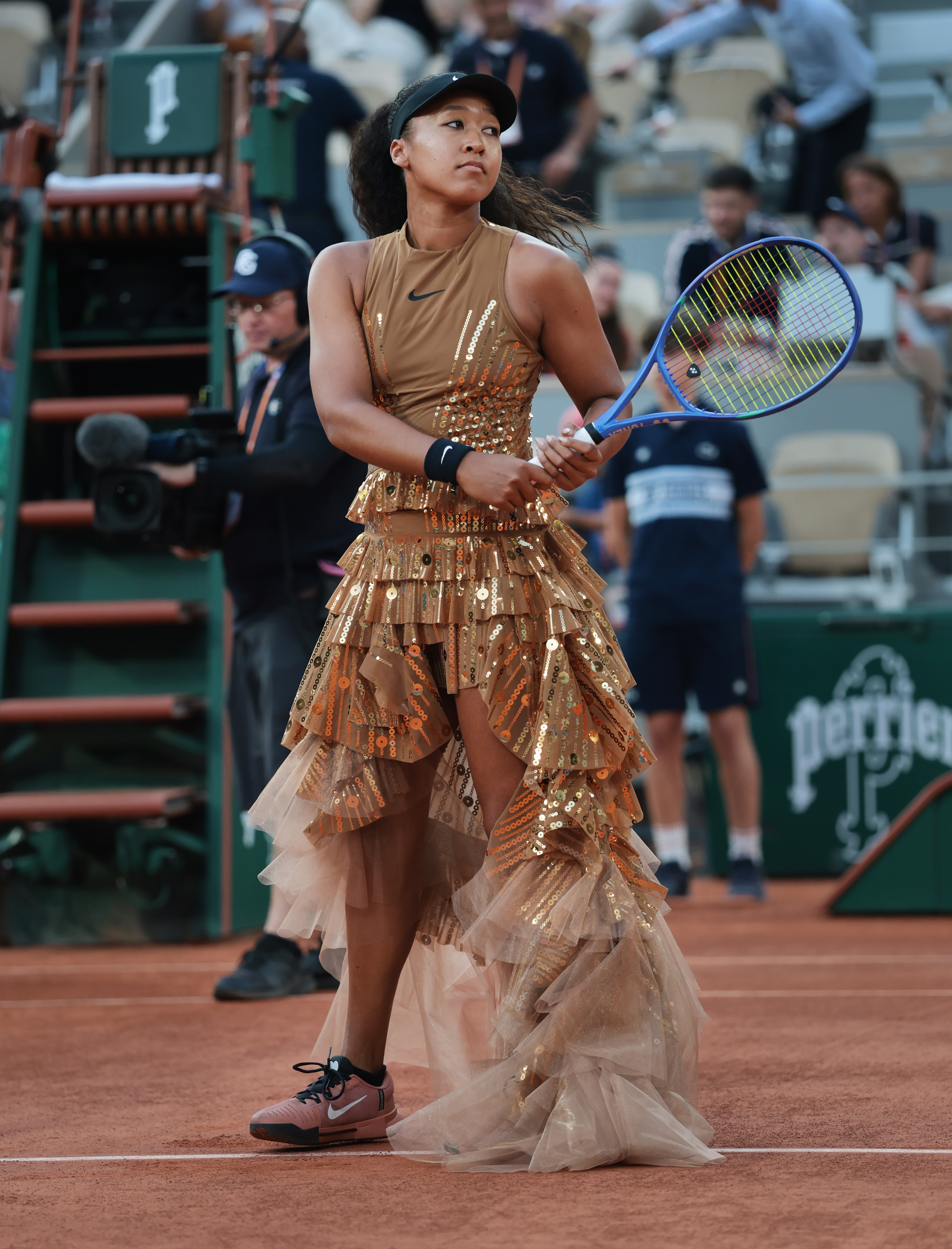 Click to open the image gallery Female tennis player standing on a clay court in a gold sequined tiered gown with a tulle train, holding a blue tennis racket.