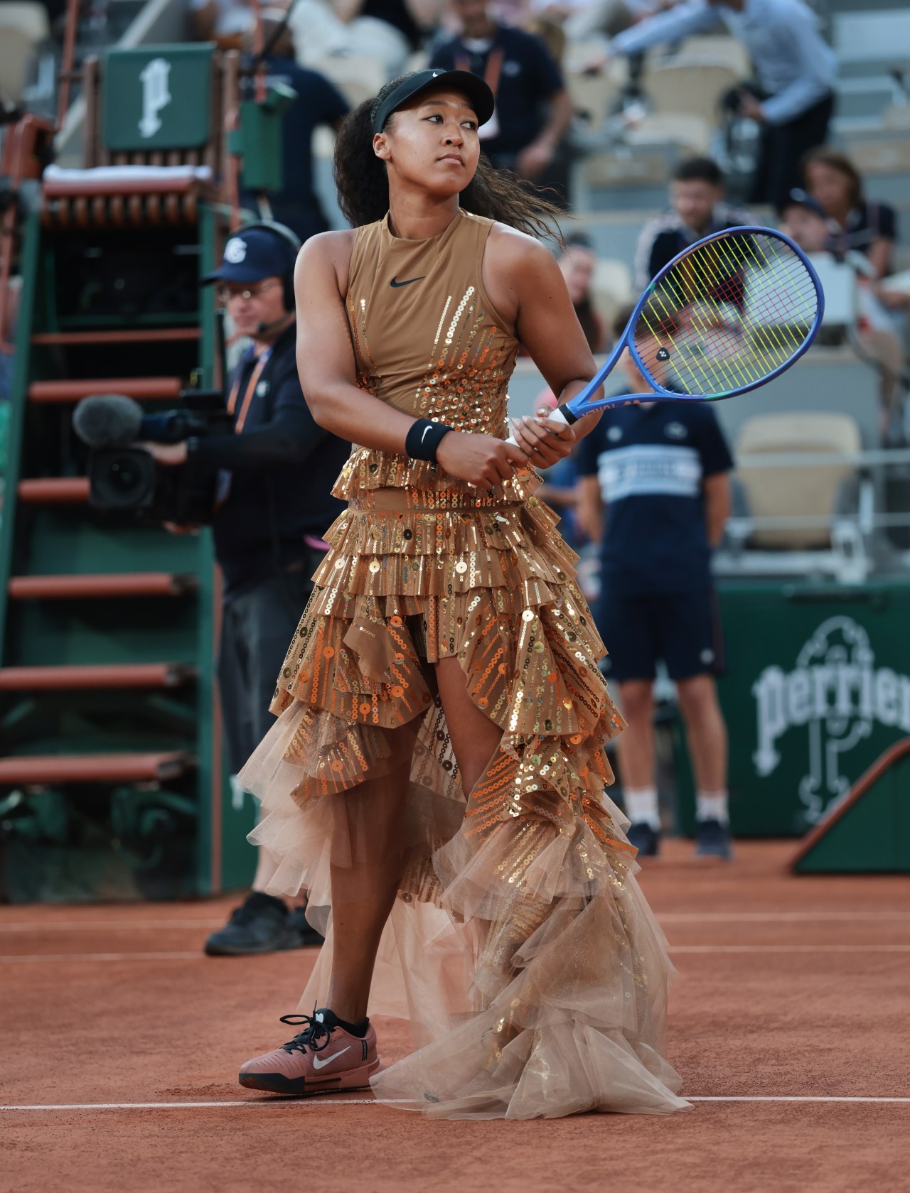 Female tennis player standing on a clay court in a gold sequined tiered gown with a tulle train, holding a blue tennis racket.