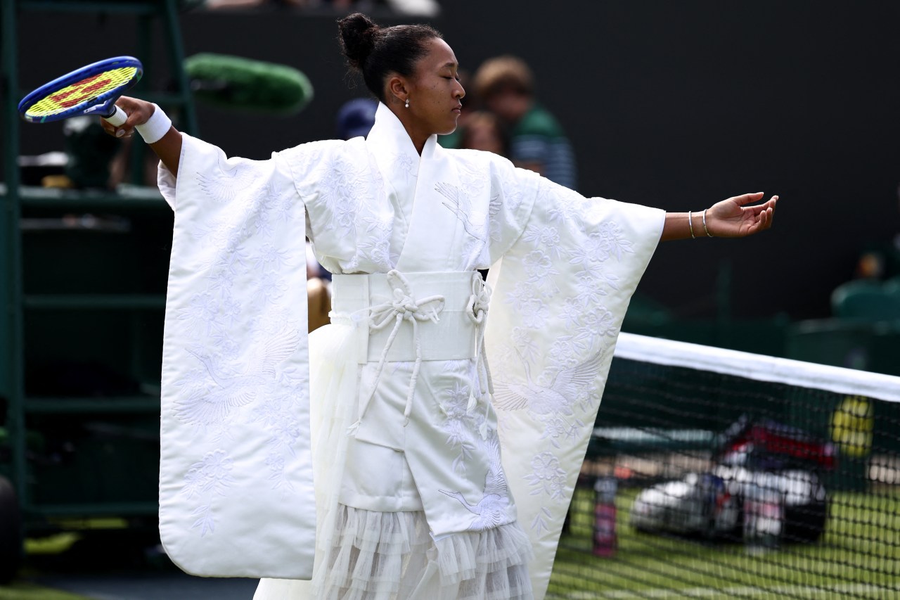 Tennis player with arms spread wide wearing a white embroidered kimono-style robe with crane motifs, standing on a grass court with a blue tennis racket in one hand.