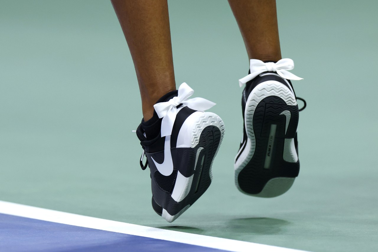 Close-up of two black-and-white Nike tennis shoes with white satin bows at the ankles, mid-air above a green hard court.