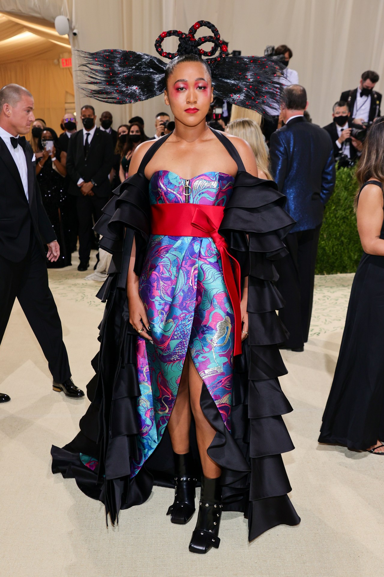 Woman in a colorful dragon-print strapless dress with a red obi belt and voluminous black ruffled cape, wearing an elaborate sculptural updo with red jewels, on a red carpet.