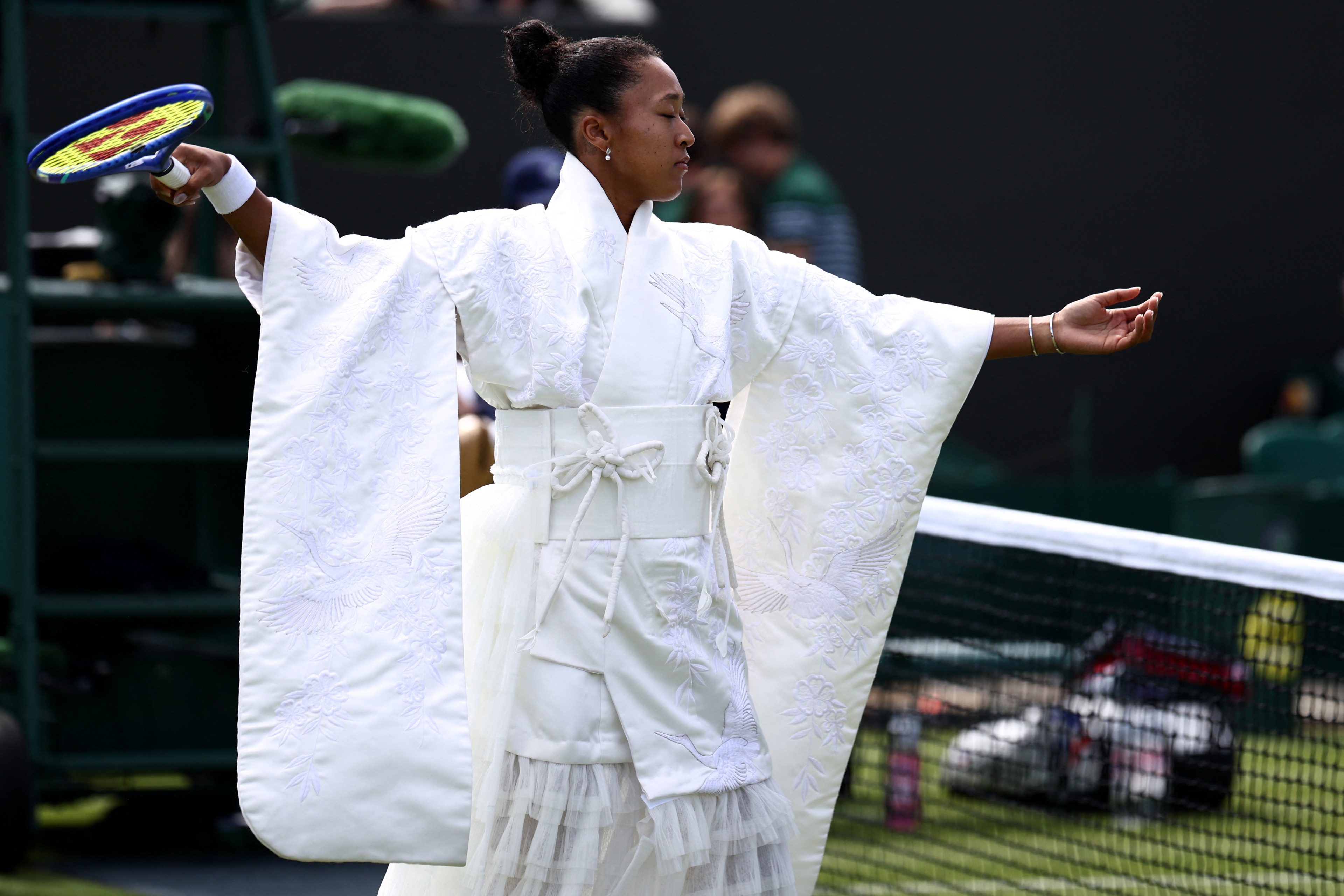 Naomi Osaka in a white embroidered kimono robe, arms outstretched, holding a tennis racket on a Wimbledon grass court.