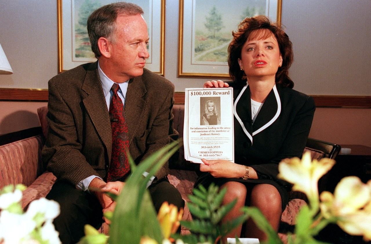 John and Patsy Ramsey sitting on a couch; Patsy holds up a $100,000 reward flyer for information in JonBenét Ramsey's murder.