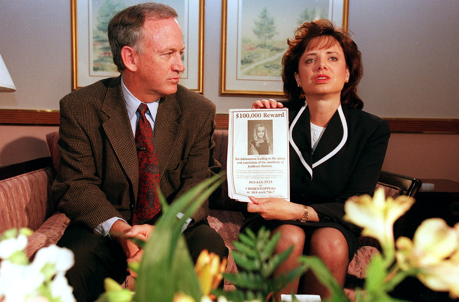 John and Patsy Ramsey sitting on a couch; Patsy holds up a $100,000 reward flyer for information in JonBenét Ramsey's murder.