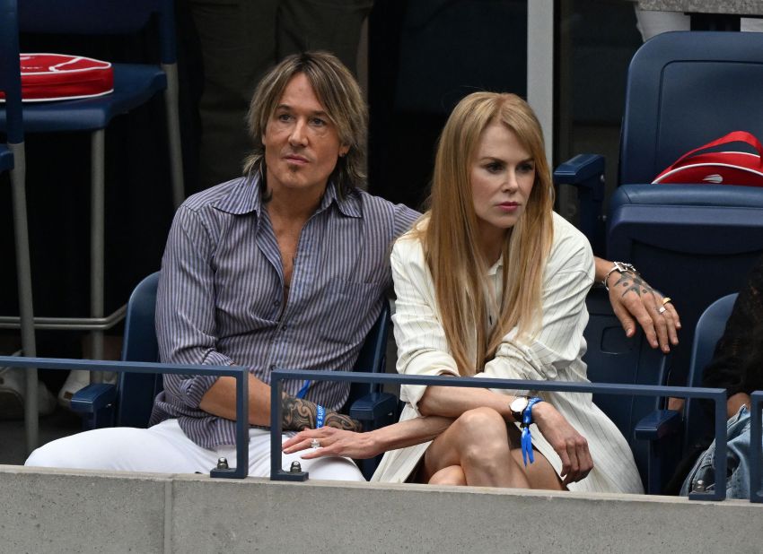 A tattooed man in a striped shirt and a blonde woman in a white outfit sit side by side in blue stadium seats at a tennis event.