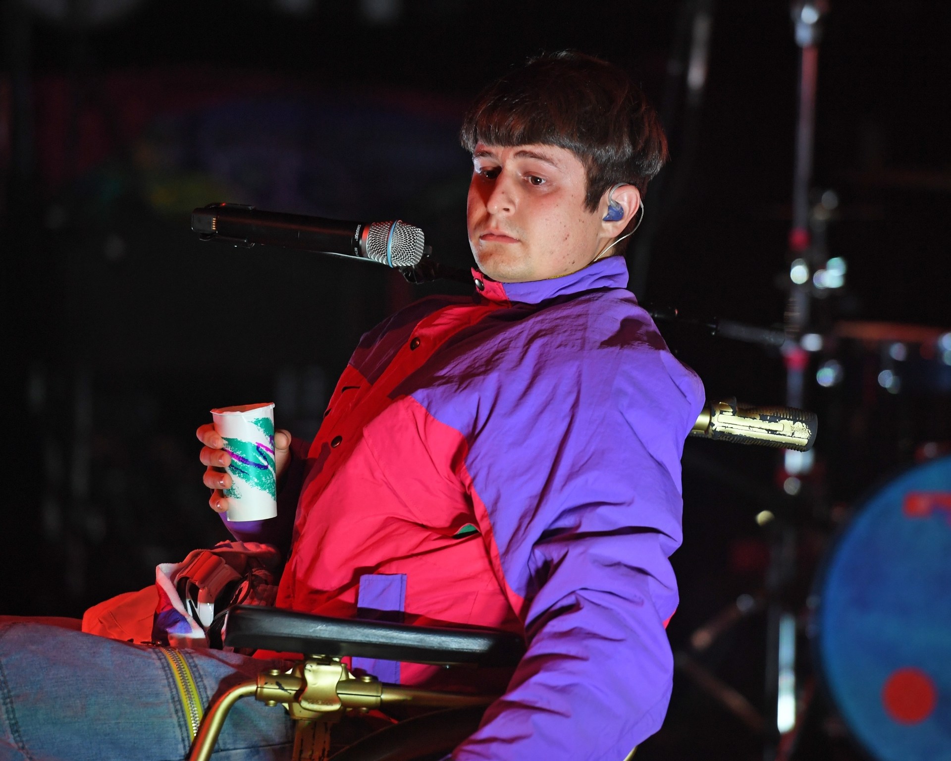 Oliver Tree seated in a wheelchair on stage, holding a paper cup, wearing a purple and red jacket with an in-ear monitor, microphone in front of him.