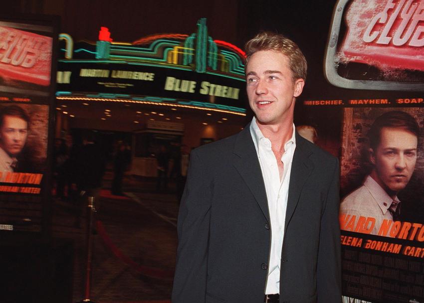 Edward Norton in a dark blazer and open white shirt standing in front of Fight Club movie posters at a theater premiere.