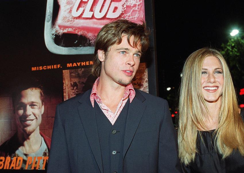 Brad Pitt in a dark blazer and pink patterned shirt stands with Jennifer Aniston, who is smiling, in front of a Fight Club movie poster.