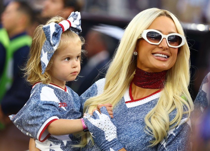 Blonde woman in white sunglasses, red rhinestone choker, and star-print jersey holds a young blonde girl in a matching outfit with a large denim bow in her hair.
