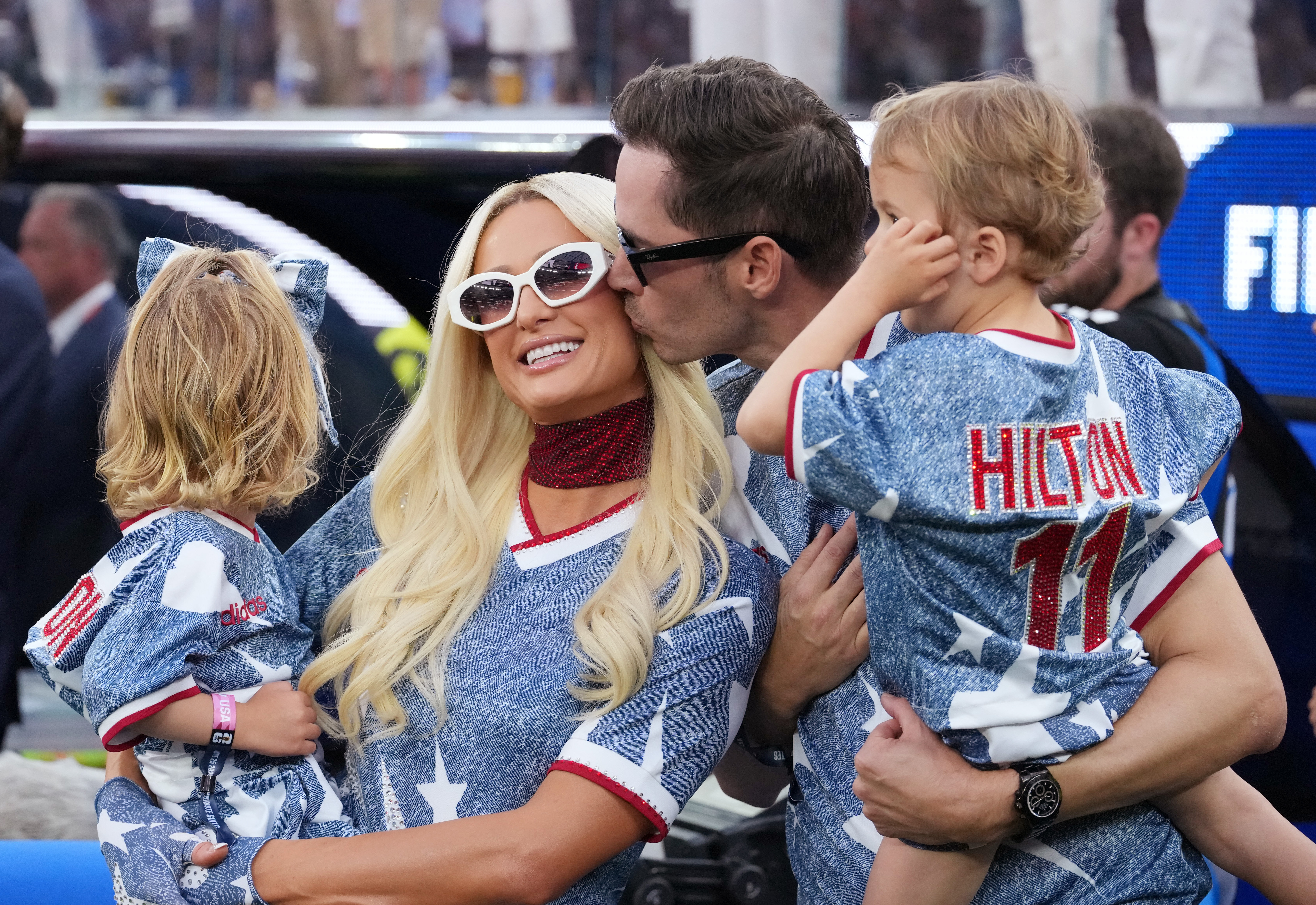 A man kisses a smiling blonde woman on the cheek while holding a toddler boy in a Hilton No. 11 jersey; a young girl with a denim bow faces away on the left.
