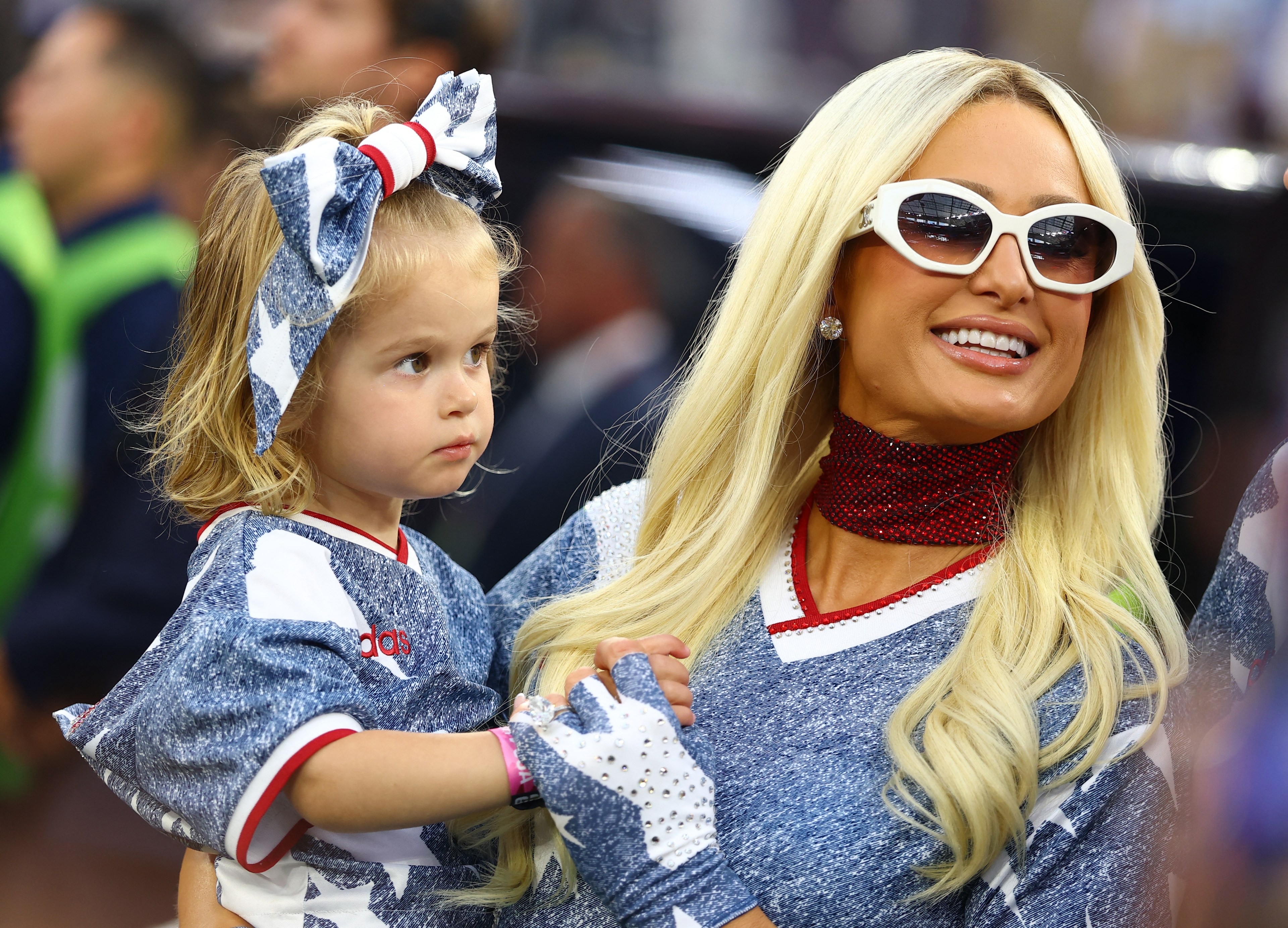 Blonde woman in white sunglasses and denim jersey holding a young blonde girl with a large bow at a soccer match.