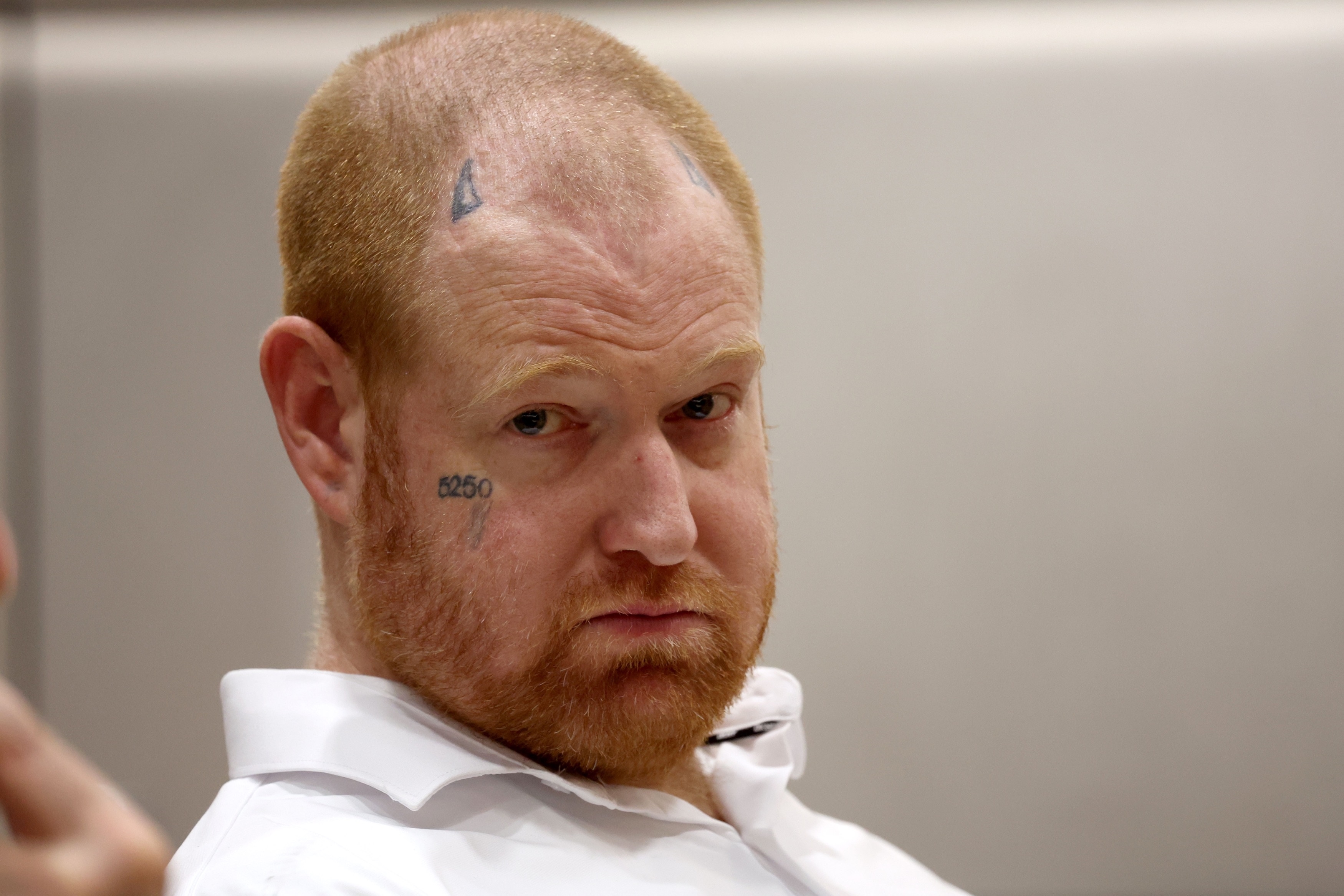 Man with red stubble, face tattoos reading "5250" on cheek and symbols on forehead, wearing a white dress shirt, looking over his shoulder.