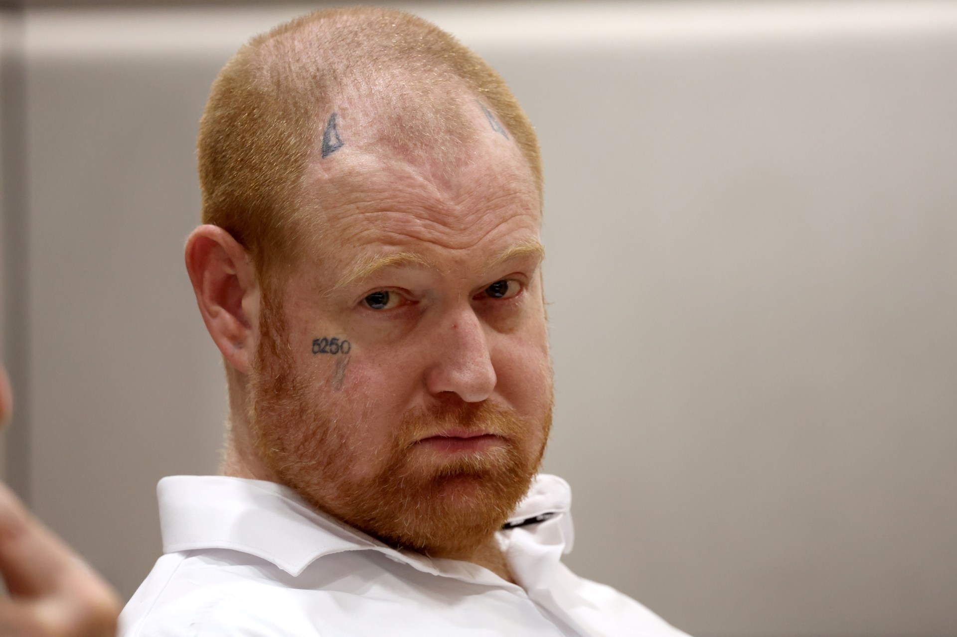 Man with red stubble, face tattoos reading "5250" on cheek and symbols on forehead, wearing a white dress shirt, looking over his shoulder.