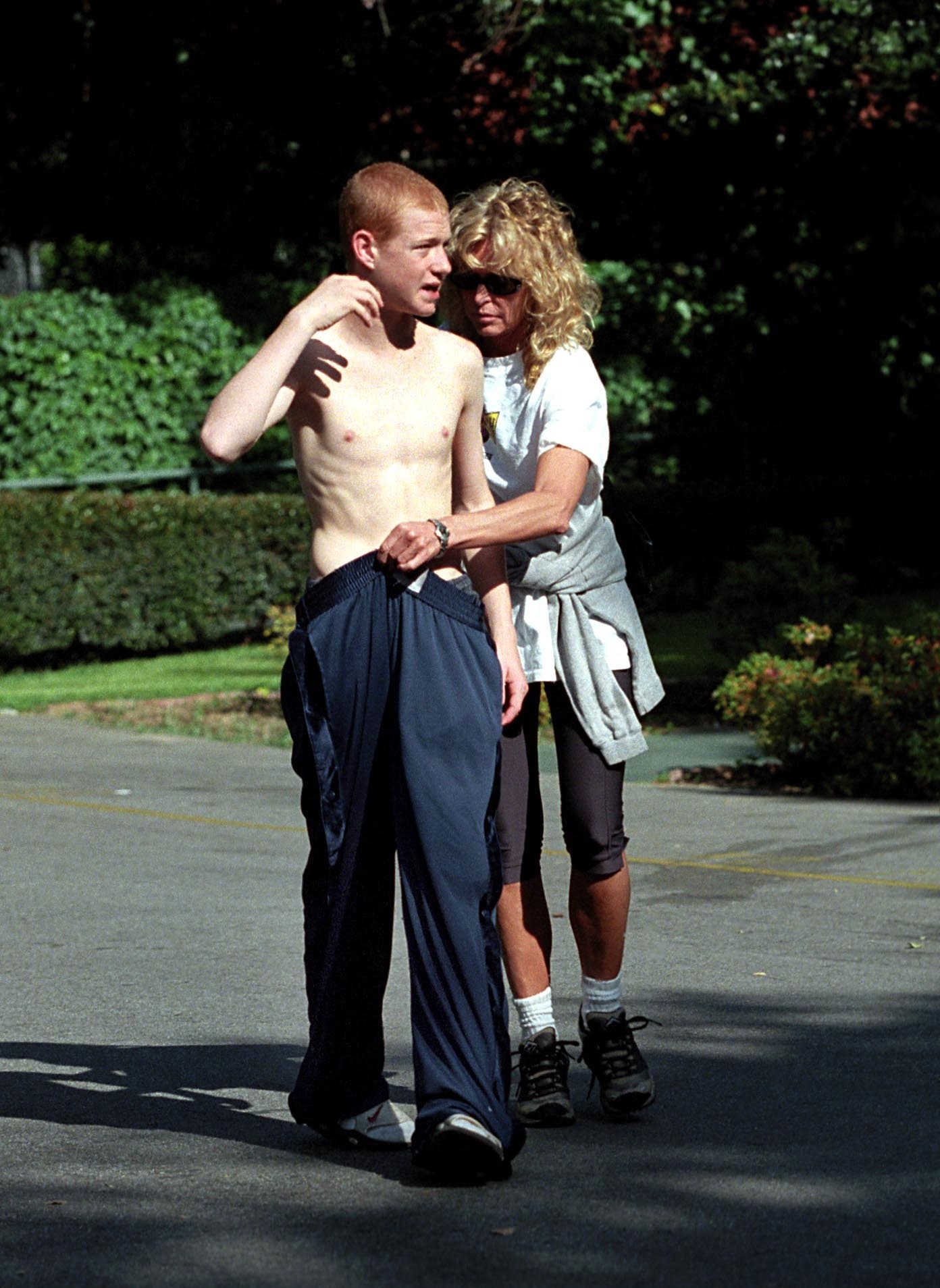 Shirtless young red-haired man in navy track pants stands outdoors while a curly-haired blond woman in a white T-shirt rests her hand on his shoulder.