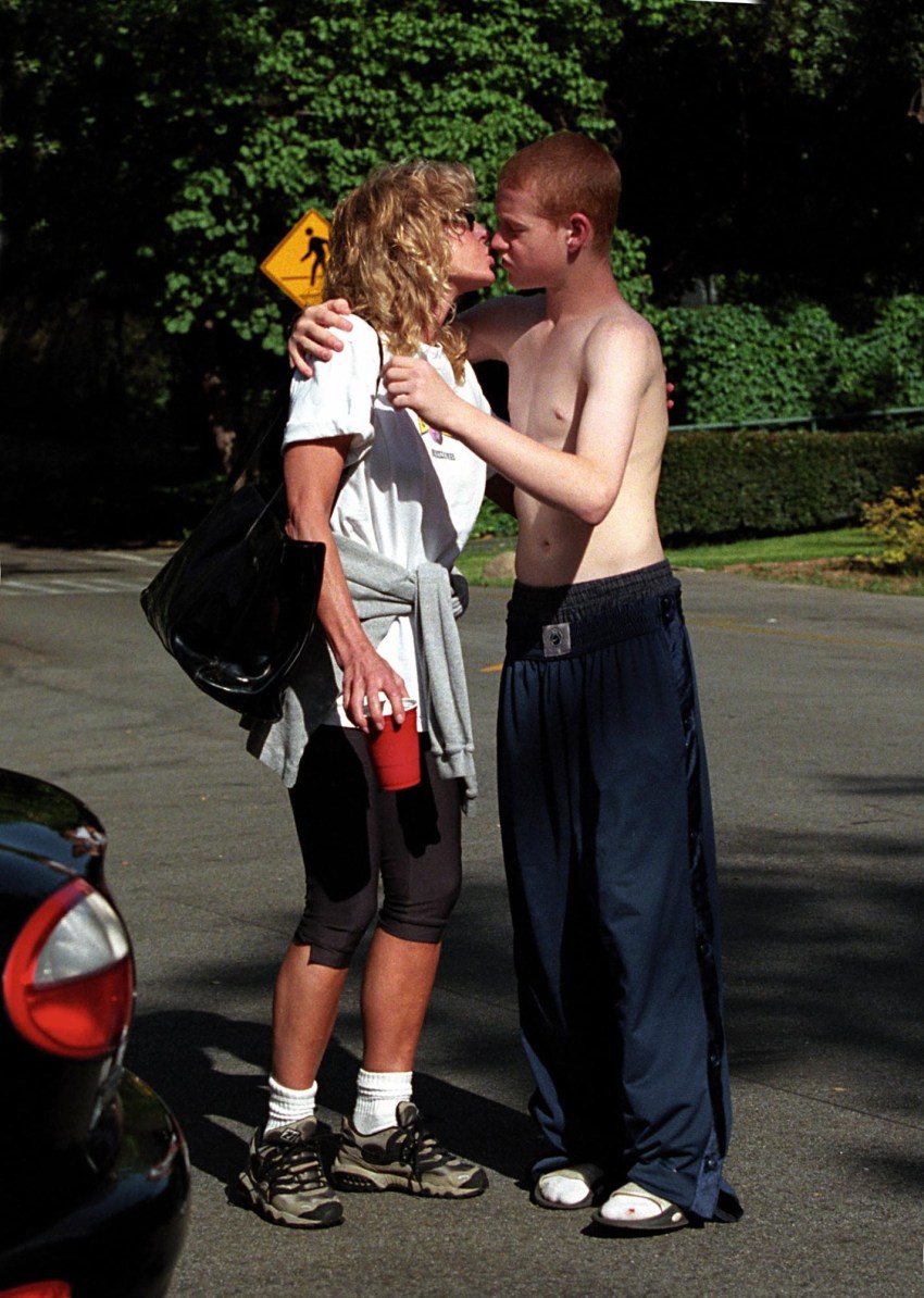 Shirtless young red-haired man in navy track pants and a blond curly-haired woman in a white T-shirt kissing on a street, with a yellow pedestrian crossing sign visible behind them.