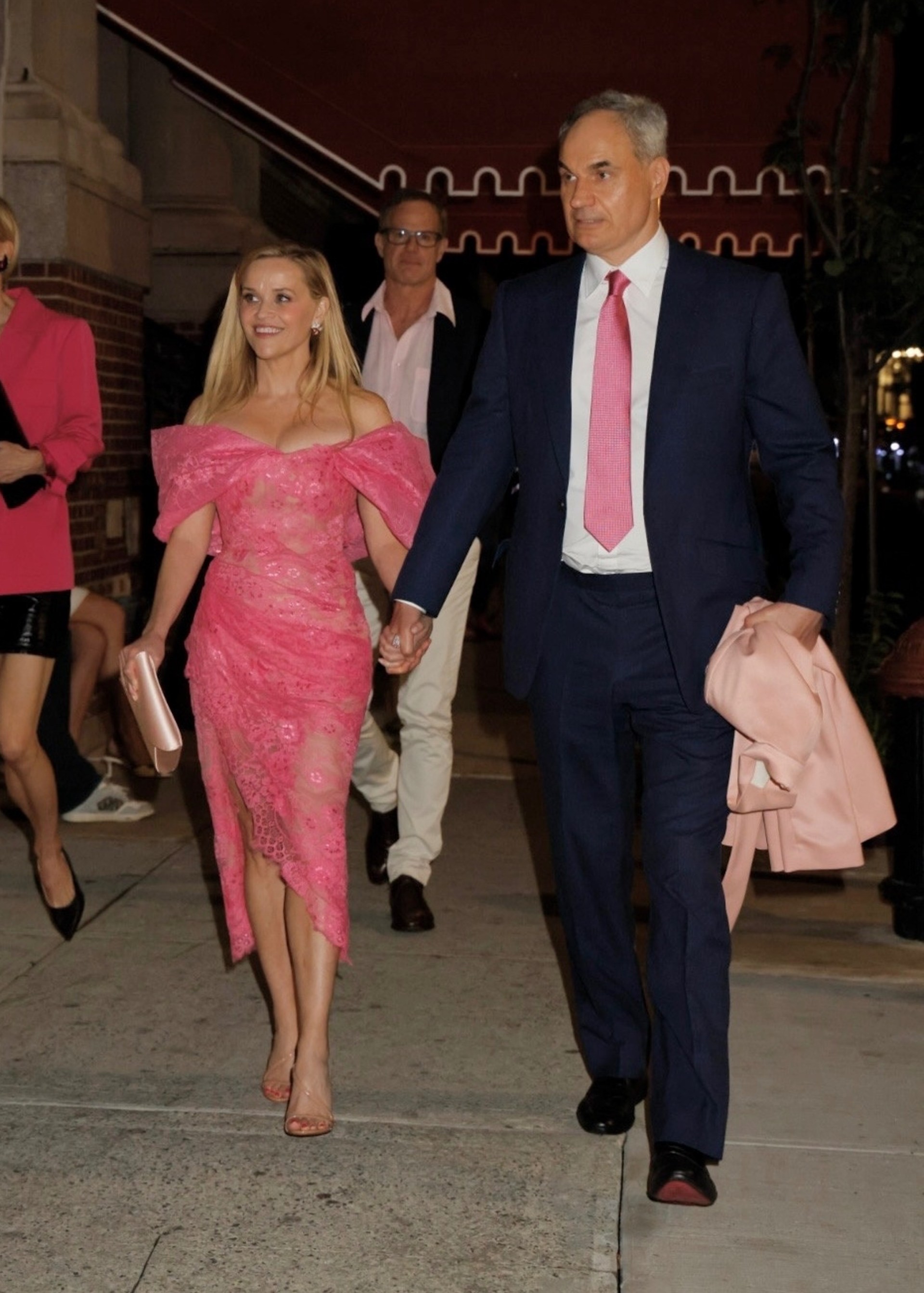 Reese Witherspoon in a pink lace off-shoulder dress holds hands with Oliver Haarmann in a navy suit and pink tie on a New York City sidewalk at night.
