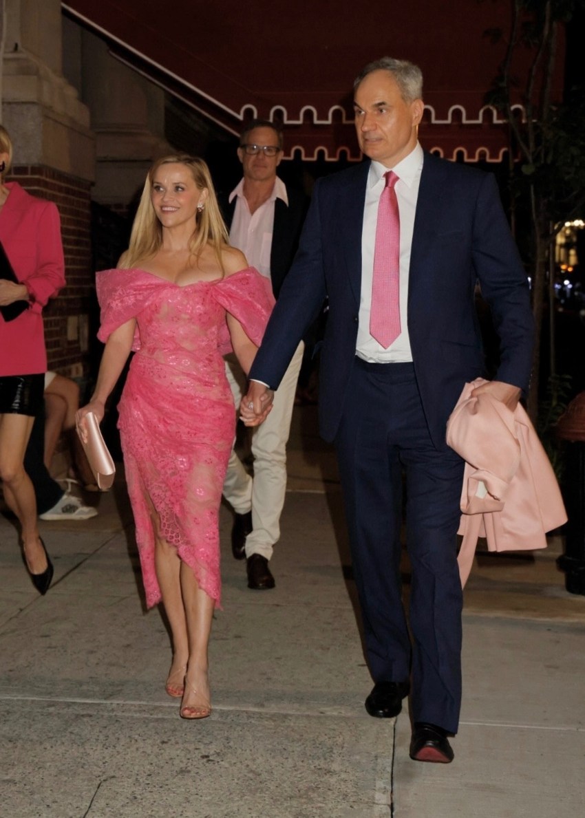 Reese Witherspoon in a pink lace off-shoulder dress holds hands with Oliver Haarmann in a navy suit and pink tie on a New York City sidewalk at night.
