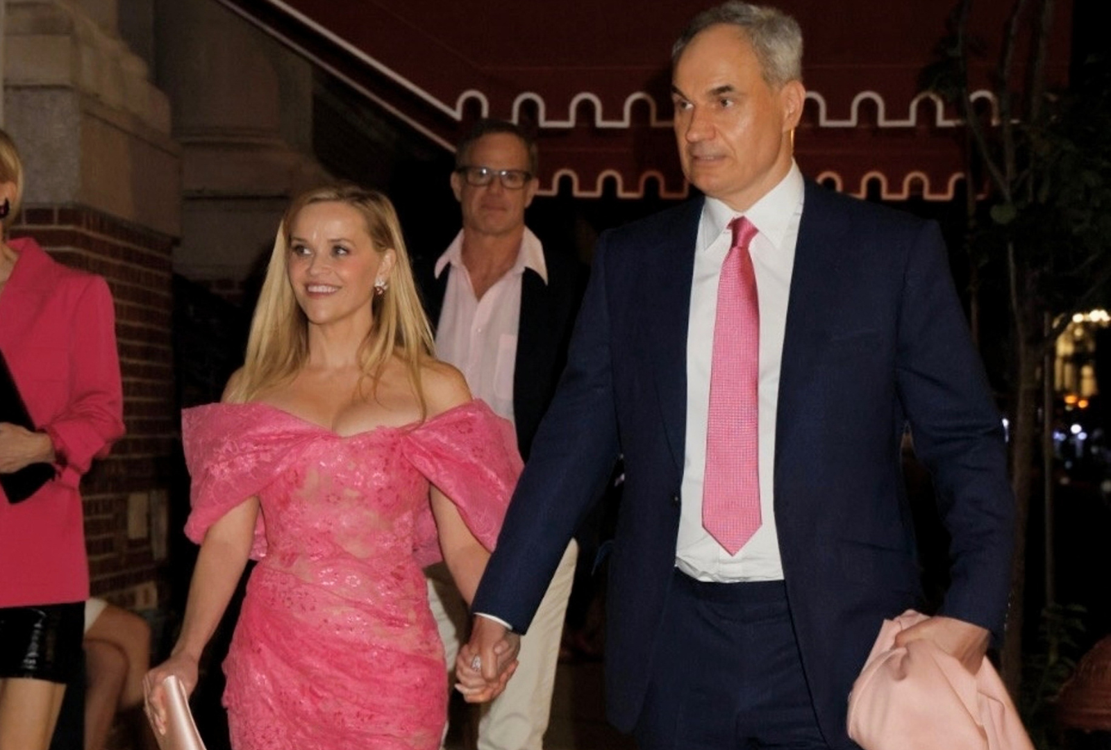 Reese Witherspoon in a pink lace off-shoulder dress holds hands with Oliver Haarmann in a navy suit and pink tie as they walk on a New York City sidewalk at night.