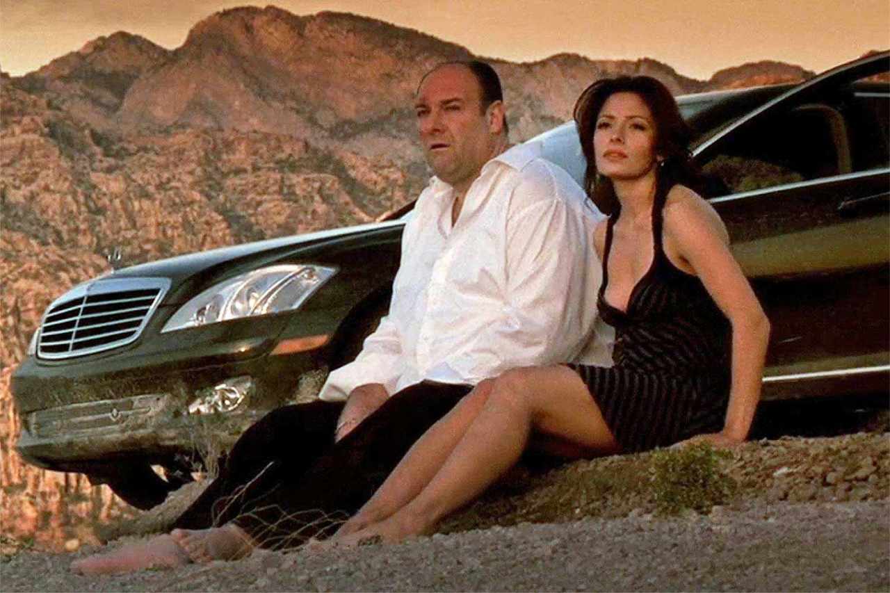A man in a white shirt and a woman in a black dress sit together in front of a black Mercedes with rocky mountains behind them.