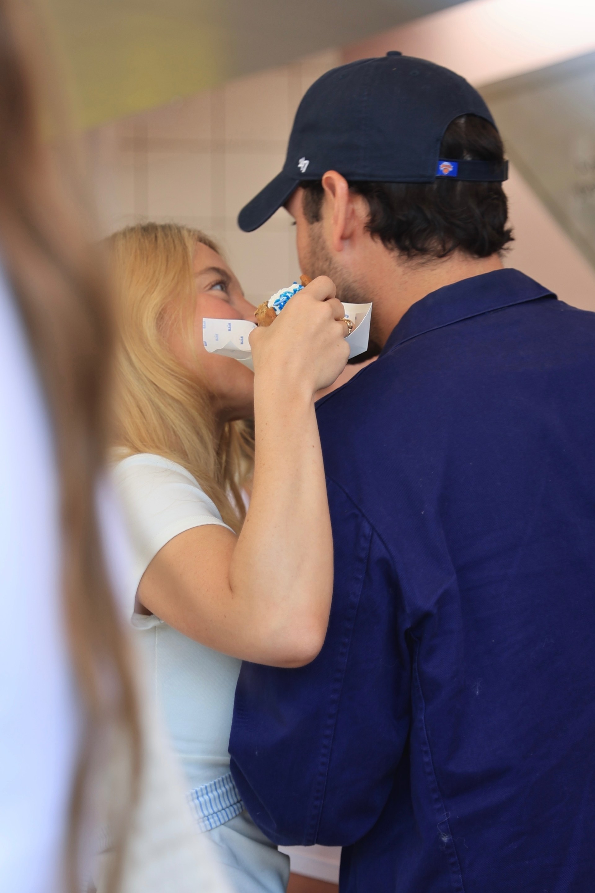 A blonde woman in a white top and a dark-haired man in a navy cap and blue shirt lean in to kiss while sharing a froyo cup.