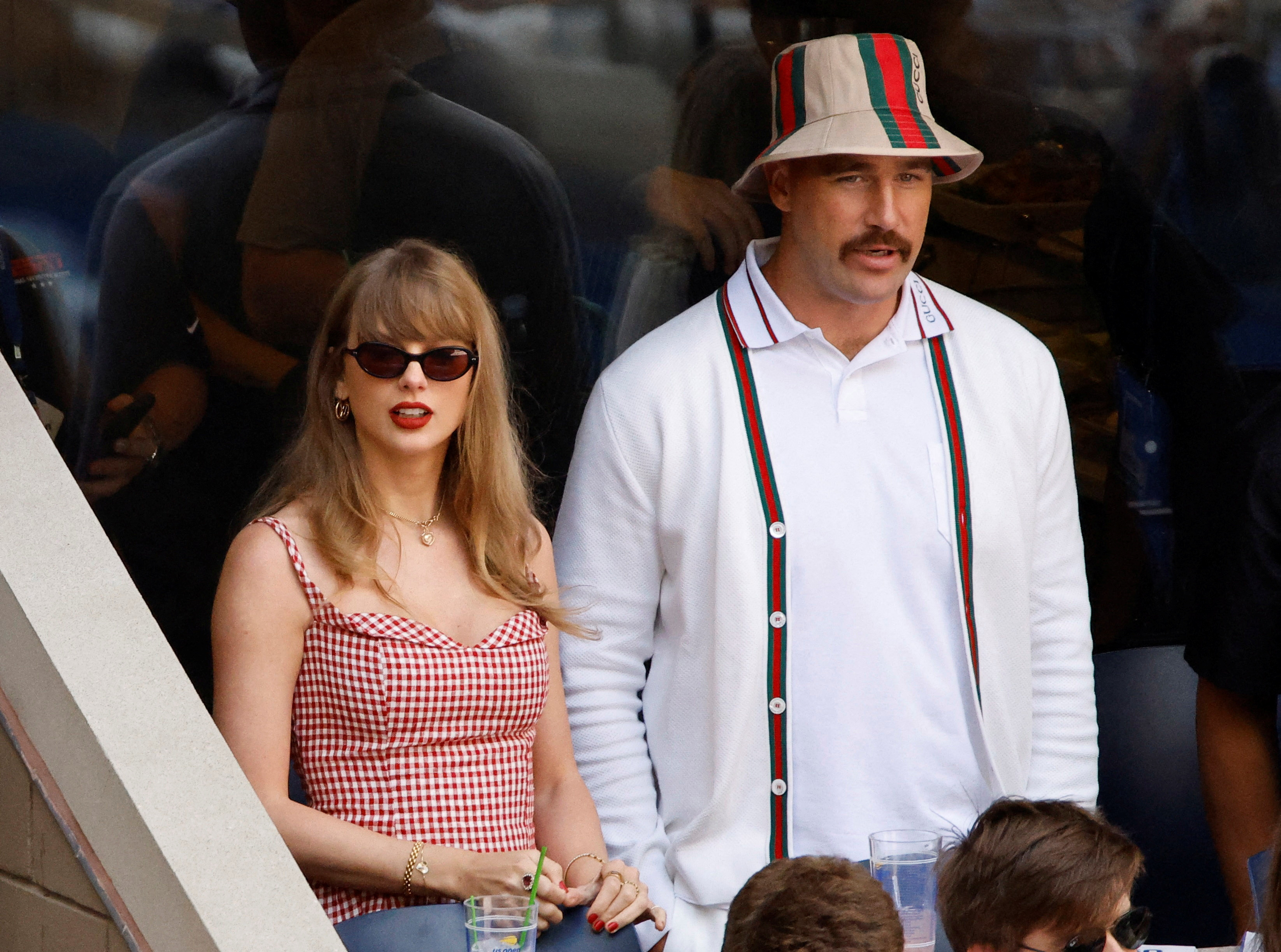 Taylor Swift in a red gingham top and sunglasses standing next to a tall man in a white Gucci cardigan and bucket hat at a tennis stadium.