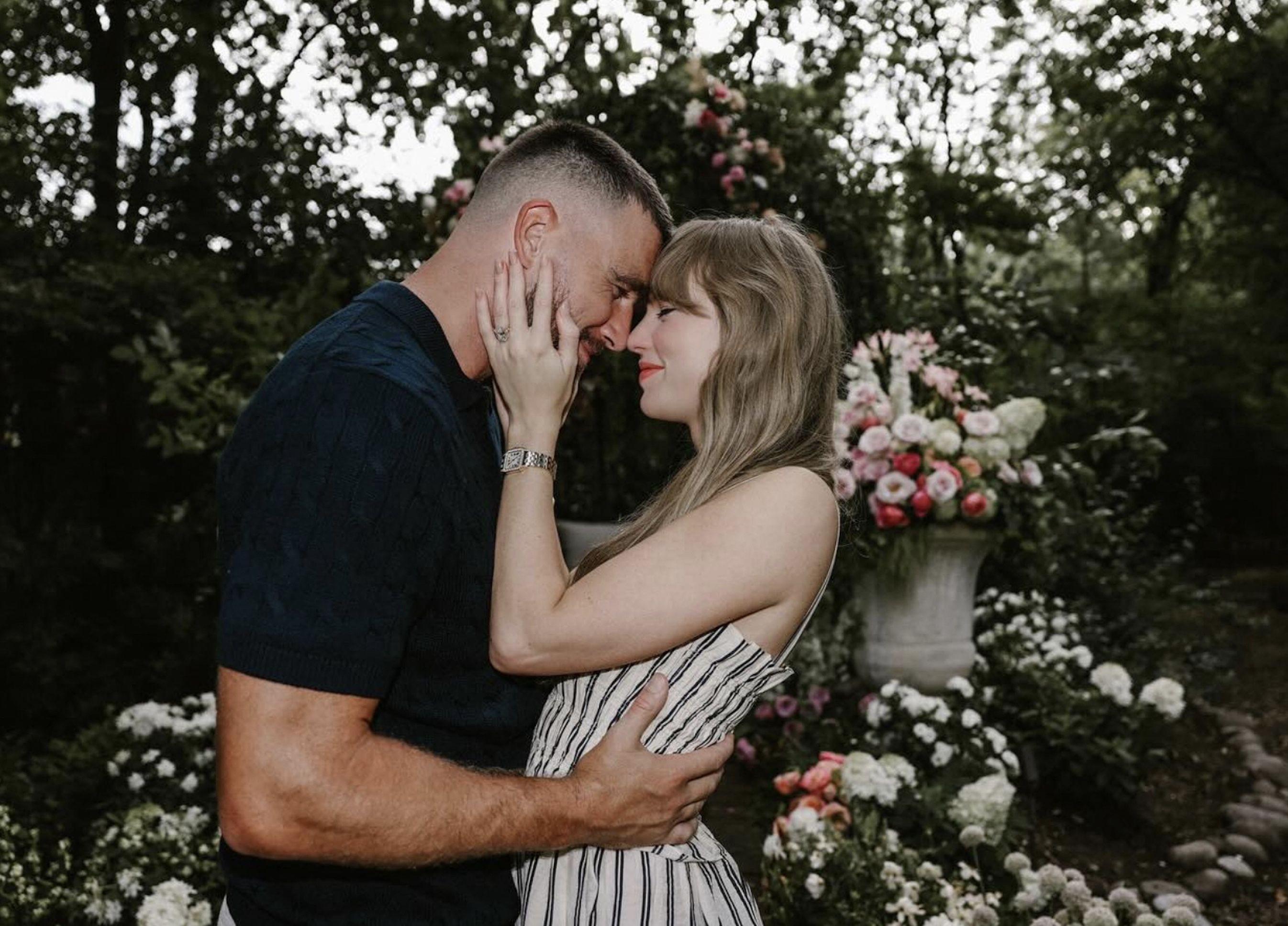 A couple touches foreheads tenderly in a lush garden surrounded by pink and white floral arrangements.
