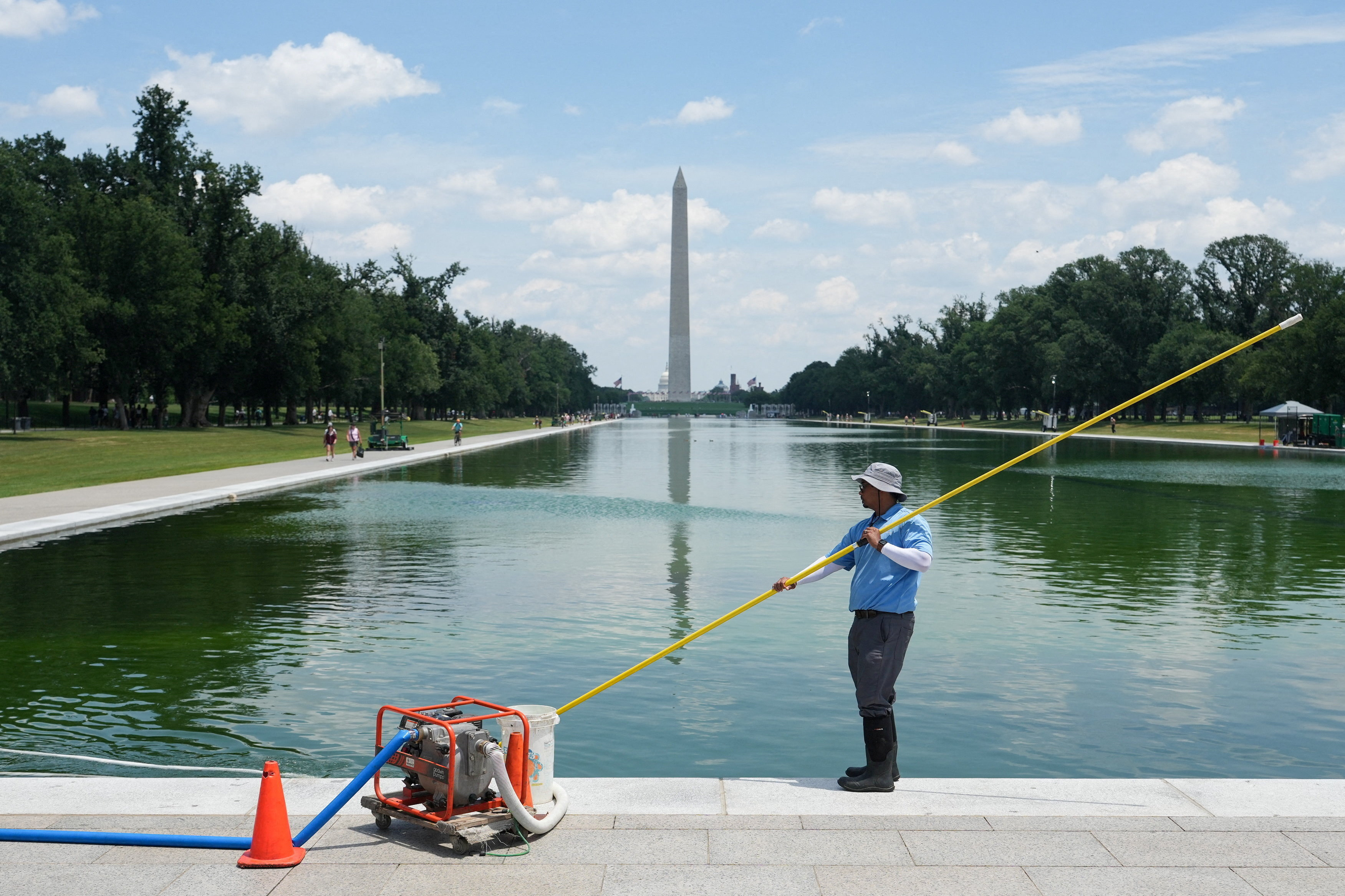 A worker in a blue shirt and hat uses a long pole to skim the Lincoln Memorial Reflecting Pool, with the Washington Monument visible in the background.