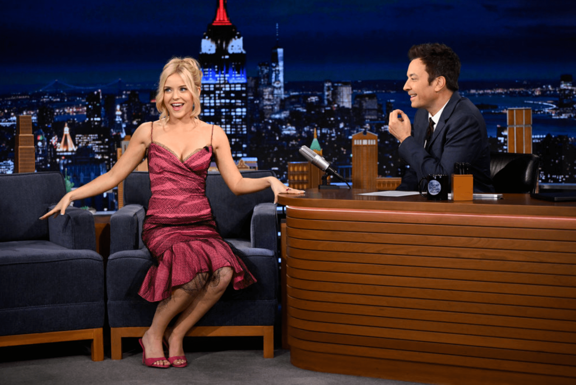 Blonde woman in a pink spaghetti-strap taffeta dress with black tulle overlay sits in the guest chair on a late-night talk show set, smiling at the host behind his desk.